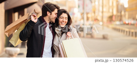A happy couple strolls down a bustling city street, laughing and carrying shopping bags filled with purchases, copy space 130574526
