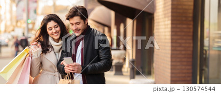 Young couple searching new boutique, using smartphone outdoors, walking with paper bags, free space 130574544