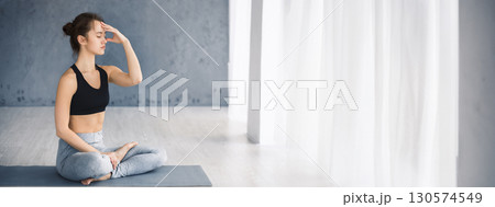 A young woman sits cross-legged on a mat, eyes closed, practicing meditation with a calm expression in a well-lit room, copy space A young woman sits cross-legged on a mat, eyes closed, practicing meditation with a calm expression in a well-lit room, copy space 130574549