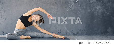 A young woman is performing a side stretch while practicing yoga on a mat in a minimalist indoor space, copy space A young woman is performing a side stretch while practicing yoga on a mat in a minimalist indoor space, copy space 130574621