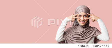 Playful arabic girl in hijab showing v-sign symbols near eyes, having fun over pink background with free space, closeup Playful arabic girl in hijab showing v-sign symbols near eyes, having fun over pink background with free space, closeup 130574635