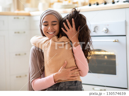 Happy Mother's Day. Little Girl Cuddling Tight Her Muslim Mom In Kitchen At Home, Expressing Love And Affection. Positive Islamic Woman In Hijab Hugging Her Child And Smiling, Closeup With Copy Space 130574639