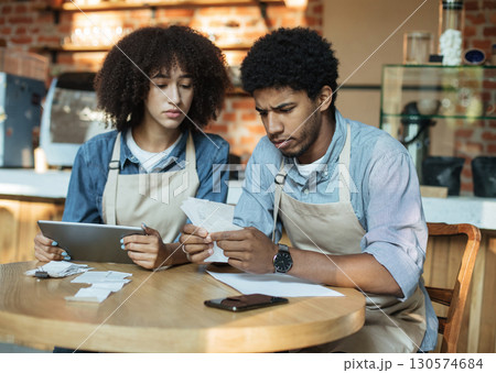 Problems in small business during covid-19 lockdown. Sad millennial african american couple owners in aprons work with accounts, and solve debt problems bookkeeping in cafe, empty space Problems in small business during covid-19 lockdown. Sad millennial african american couple owners in aprons work with accounts, and solve debt problems bookkeeping in cafe, empty space 130574684