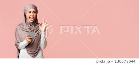 Cool islamic app. Positive muslim girl in hijab winking and showing ok gesture while using smartphone, standing over pink background with copy space Cool islamic app. Positive muslim girl in hijab winking and showing ok gesture while using smartphone, standing over pink background with copy space 130575044