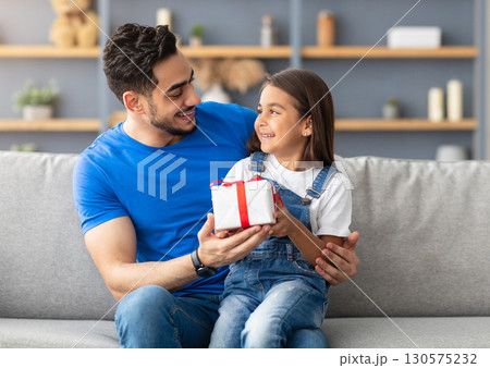 This Present Is For You. Portrait of loving girl sitting on dad's lap and greeting him with father's day or birthday, holding wrapped gift box, happy family celebrating holiday together at home This Present Is For You. Portrait of loving girl sitting on dad's lap and greeting him with father's day or birthday, holding wrapped gift box, happy family celebrating holiday together at home 130575232