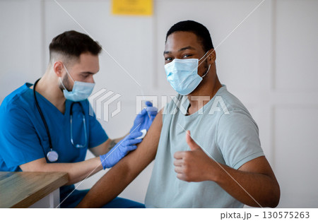 African American man in antiviral mask gesturing thumb up during coronavirus vaccination, approving of covid-19 immunization. Young doctor giving vaccine injection to male patient 130575263