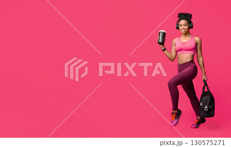 Excited african fitness girl with earphones, bag and shaker happily running to gym over red background 130575271