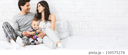 A joyful family sits on a cozy bed, engaging with their baby who is smiling and playing with colorful toys. The parents share a loving glance while enjoying this quality time together in bright room 130575278