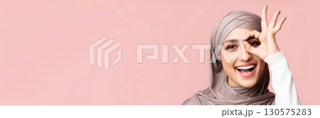 Cheerful arabic lady in hijab showing ok gesture, holding hand near eye, looking through fingers at camera, pink background with empty space, closeup 130575283