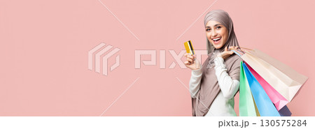 Happy customer. Joyful muslim girl in hijab with colorful shopping bags and credit card over pink studio background and looking at camera, empty space 130575284