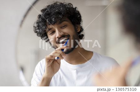 Oral care concept. Young indian man cleaning teeth with toothbrush, smiling to his reflection in mirror, doing toothcare hygiene routine in the morning in bathroom Oral care concept. Young indian man cleaning teeth with toothbrush, smiling to his reflection in mirror, doing toothcare hygiene routine in the morning in bathroom 130575310