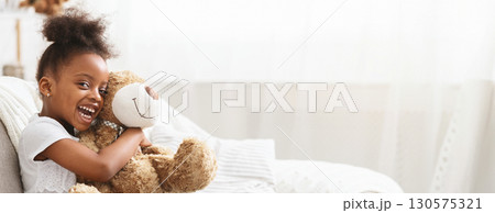 African American young girl with curly hair looks delighted as she hugs a soft teddy bear in a bright, cozy living room. Sunlight filters through sheer curtains, creating a warm atmosphere. 130575321