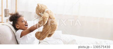 A cheerful young black girl holds up a plush teddy bear with excitement, showcasing her joy. She sits comfortably on a couch, copy space A cheerful young black girl holds up a plush teddy bear with excitement, showcasing her joy. She sits comfortably on a couch, copy space 130575430