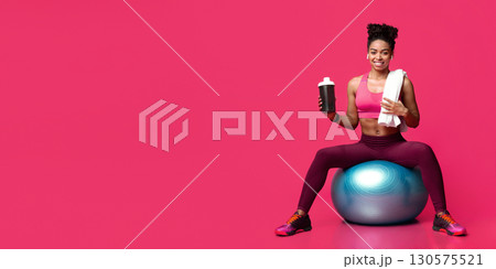 Smiling athletic black girl in sportswear recommending sports food, sitting on fitness ball, holding shaker, sports food Smiling athletic black girl in sportswear recommending sports food, sitting on fitness ball, holding shaker, sports food 130575521