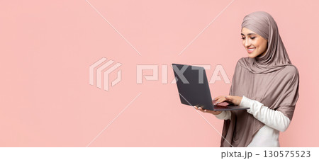 Freelance and online education for muslim women concept. Smiling arabian woman in hijab holding and typing on laptop over pink background, copy space Freelance and online education for muslim women concept. Smiling arabian woman in hijab holding and typing on laptop over pink background, copy space 130575523