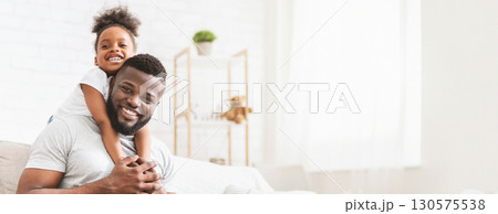 African American father and daughter are enjoying a special bonding moment in their cozy living room. The girl, smiling widely, is playfully hugging her dad from behind while he sits comfortably 130575538