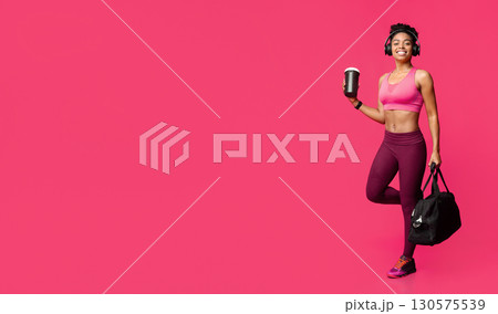 Excited african fitness girl with earphones, bag and shaker going to gym, red background 130575539