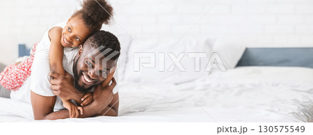 Black father enjoys a playful moment with his daughter on a cozy bed, filled with laughter and joy. They share a heartfelt connection, creating a loving atmosphere while engaging in fun activities Black father enjoys a playful moment with his daughter on a cozy bed, filled with laughter and joy. They share a heartfelt connection, creating a loving atmosphere while engaging in fun activities 130575549