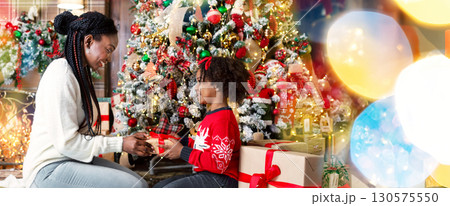 Merry Christmas. African american mother and daughter exchanging gifts sitting under Xmas tree at home, side view 130575550