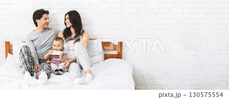A joyful family is enjoying quality time together while sitting on a bed. The parents, dressed casually in comfortable attire, are smiling at each other, while their adorable baby sits between them A joyful family is enjoying quality time together while sitting on a bed. The parents, dressed casually in comfortable attire, are smiling at each other, while their adorable baby sits between them 130575554