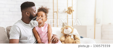 Black father embraces his young daughter on a comfortable couch, both smiling happily. The cozy living room features soft decor and a stuffed teddy bear nearby, copy space 130575559