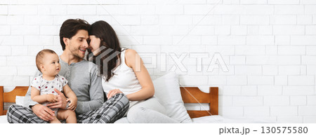 A joyful family shares affectionate moments as they sit on a comfortable bed. The mother and father gaze lovingly at each other while holding their baby, creating a warm and loving atmosphere 130575580