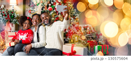 Happy african american family of three taking selfie near Christmas tree at home, sitting on floor close to fireplace in decorated living room 130575604