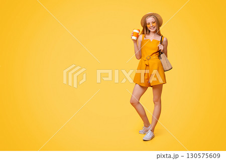 Summer Girl. Stylish Young Woman In Hat. Sunglasses And Jumpsuit Enjoying Her Takeaway Drink Over Yellow Studio Background, Copy Space 130575609