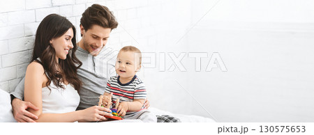 A joyful family engages in playful activities together in a bright living room. The parents sit on a comfortable bed with their toddler, who eagerly plays with colorful toys, copy space 130575653