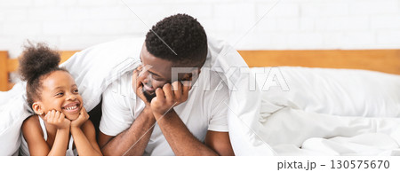 Black father and his young daughter share a joyful moment in their bed, laughing and enjoying each other's company. They are wrapped in a cozy blanket, with bright smiles, copy space 130575670