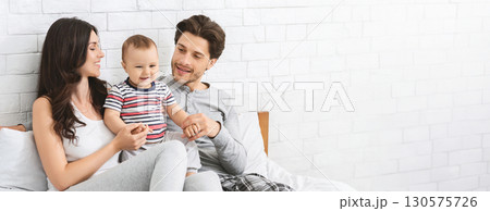 A joyful young couple sits on a bed, engaging with their cheerful baby. The parents smile at each other and the child, creating a warm atmosphere. Soft light fills the room, copy space 130575726