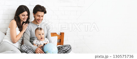 A mother and father sit together on a comfortable bed, actively engaging their young child who is fascinated by a globe. The parents share smiles and joy as they explore the world, copy space A mother and father sit together on a comfortable bed, actively engaging their young child who is fascinated by a globe. The parents share smiles and joy as they explore the world, copy space 130575736