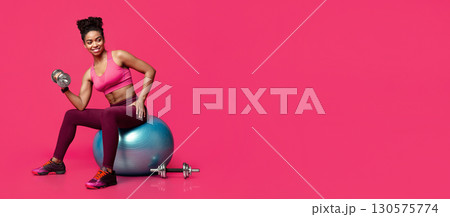 Black fitness girl exercising with dumbbells, sitting on fitness ball over red studio background, free space Black fitness girl exercising with dumbbells, sitting on fitness ball over red studio background, free space 130575774