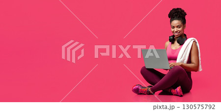Smiling sporty athletic afro woman using laptop over red background, hanging towel and earphones, panorama with free space Smiling sporty athletic afro woman using laptop over red background, hanging towel and earphones, panorama with free space 130575792