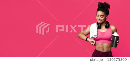 Healthy lifestyle. African fitness girl with towel on her shoulder and wireless headset holding bottle with protein, looking at sport watch Healthy lifestyle. African fitness girl with towel on her shoulder and wireless headset holding bottle with protein, looking at sport watch 130575809