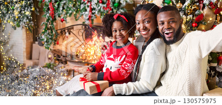 Happy afro parents taking selfie with their cute little daughter near Christmas tree, sitting on floor in decorated living room Happy afro parents taking selfie with their cute little daughter near Christmas tree, sitting on floor in decorated living room 130575975