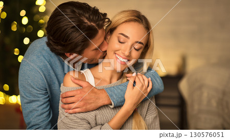 Man kissing neck of stunning woman, sitting near Christmas tree and enjoying time together 130576051