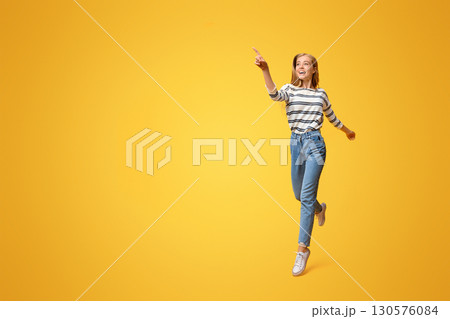 Look there. Joyful girl jumping and pointing on copy space, showing something on yellow background Look there. Joyful girl jumping and pointing on copy space, showing something on yellow background 130576084