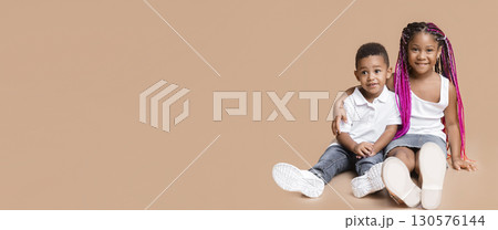 Happy childhood. Portrait of cute little afro kids, brother and sister sitting on floor and cuddling, yellow studio background with free space 130576144