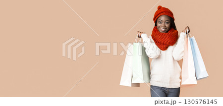 A joyful young African American woman stands smiling while holding several colorful shopping bags. She is dressed warmly in a cozy sweater and a bright red winter hat, copy space A joyful young African American woman stands smiling while holding several colorful shopping bags. She is dressed warmly in a cozy sweater and a bright red winter hat, copy space 130576145