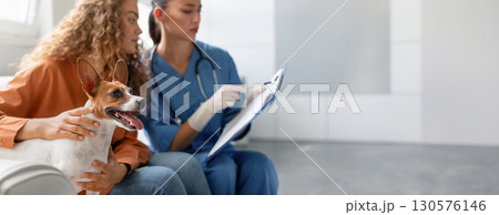Attentive vet takes clinical notes while a pet owner lovingly strokes her happy Jack Russell Terrier at a routine vet appointment in a bright clinic, copy space Attentive vet takes clinical notes while a pet owner lovingly strokes her happy Jack Russell Terrier at a routine vet appointment in a bright clinic, copy space 130576146