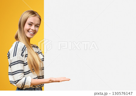 Look here. Beautiful teenage girl pointing at white blank placard with open palm, orange background Look here. Beautiful teenage girl pointing at white blank placard with open palm, orange background 130576147