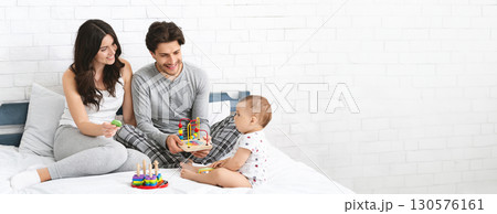 A young couple enjoys quality time together in a bright room while engaging their baby with colorful toys on a comfortable bed. The atmosphere is filled with warmth and joy, copy space 130576161