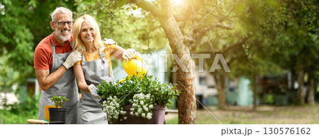 Happy Mature Gardeners Couple In Aprons Tending To Plants In Backyard, Loving Senior Spouses Watering Flowers And Smiling At Camera, Older Man And Woman Enjoying Retirement Leisure, Copy Space 130576162