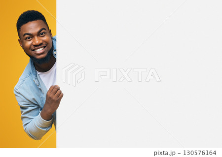Young african-american man looking out of white blank placard ready for your text or product, orange background 130576164