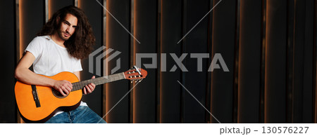 Talented Hispanic Musician Man Playing Acoustic Guitar, Touching Strings Of Musical Instrument And Looking At Camera Posing In Dark Studio Indoor, Standing Near Empty Space For Advertisement 130576227