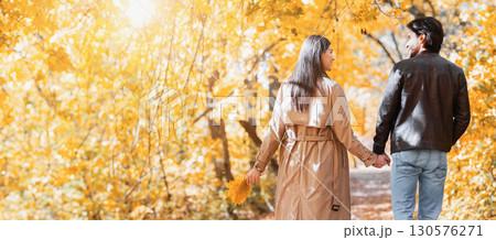Rear view of young man and woman lovers holding hands and walking by golden forest, sun flare, copy space 130576271