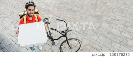 Order delivery. Smiling young man with beard, in helmet, shows box of pizza to peephole, near bike on street, free space Order delivery. Smiling young man with beard, in helmet, shows box of pizza to peephole, near bike on street, free space 130576280