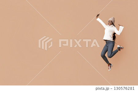 Lifeful black woman in warm hat enjoying winter over beige studio background, jumping up, copy space Lifeful black woman in warm hat enjoying winter over beige studio background, jumping up, copy space 130576281