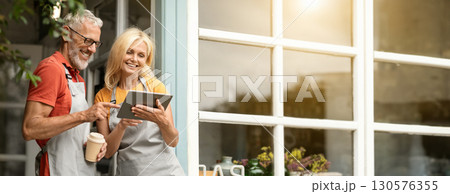 Modern Technologies. Smiling Mature Farmers Couple In Aprons Using Digital Tablet Together And Drinking Coffee While Standing At Terrace Near Country House, Happy Spouses Browsing Internet, Copy Space 130576355